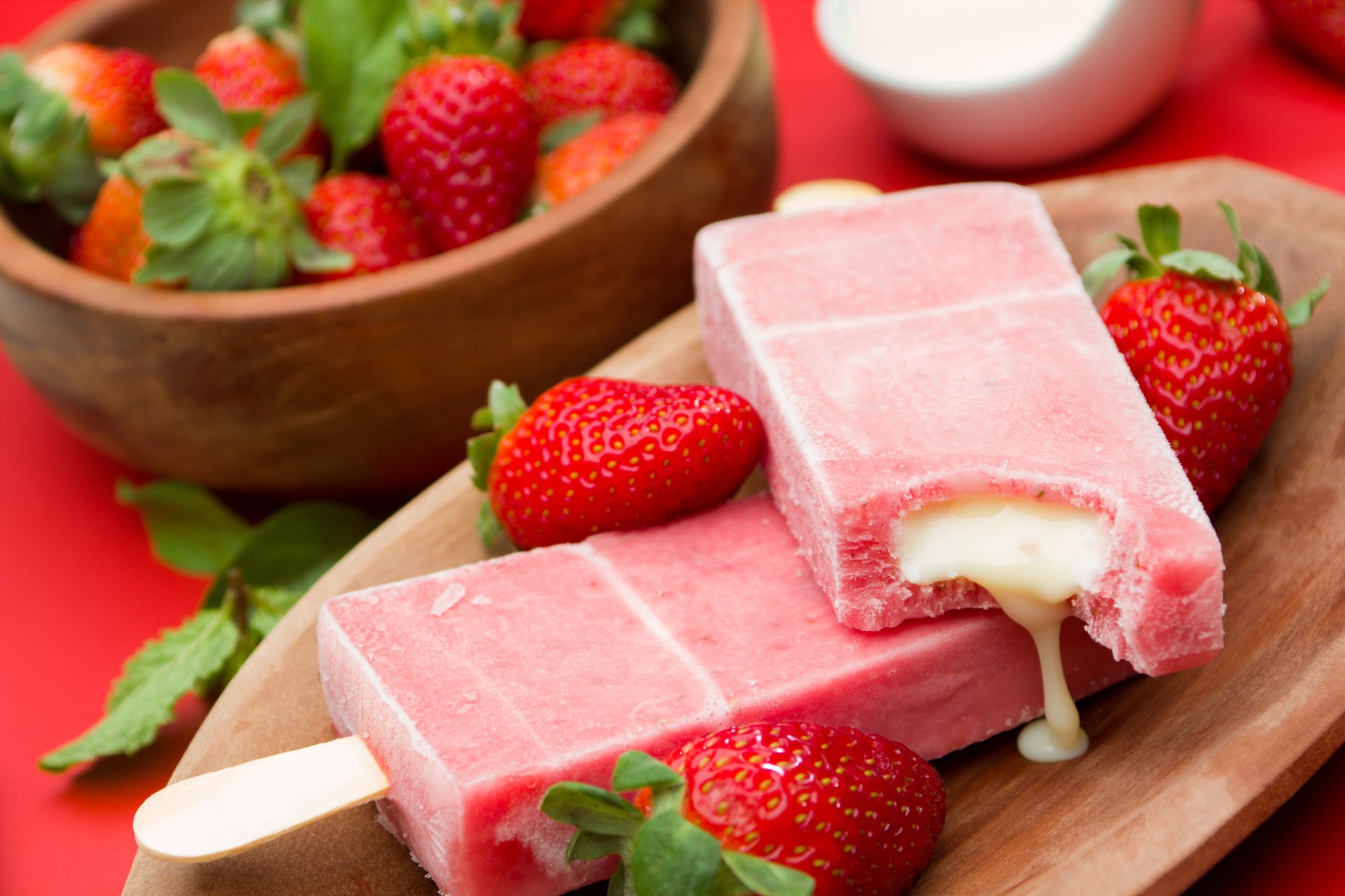 A vibrant summer scene featuring strawberry ice cream bars with gooey centers, surrounded by fresh strawberries in a wooden bowl, set against a bright red background.