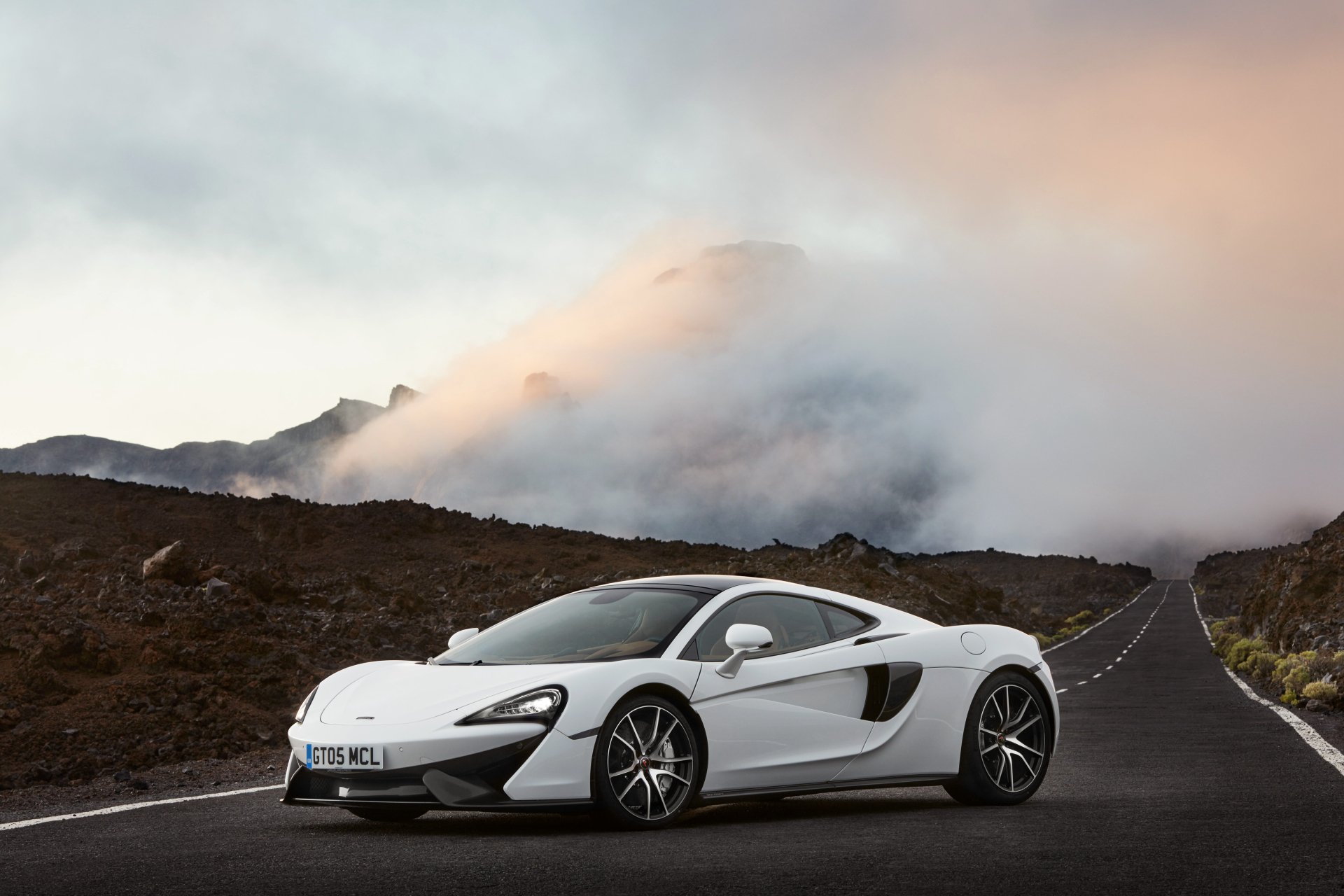 White McLaren 570GT supercar parked on a mountain road with misty hills in the background, showcased in a 4K Ultra HD desktop wallpaper.