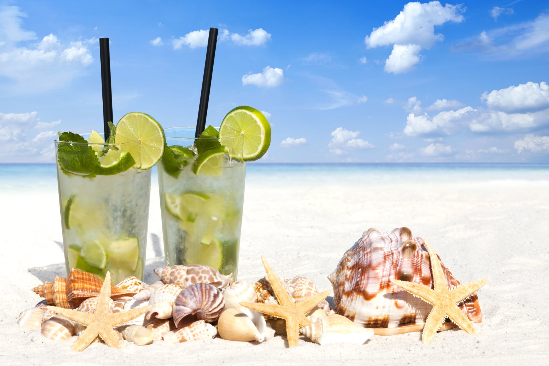 Two refreshing cocktails with lime and mint sit on a sandy beach surrounded by shells and starfish, under a clear blue sky on a bright summer day.