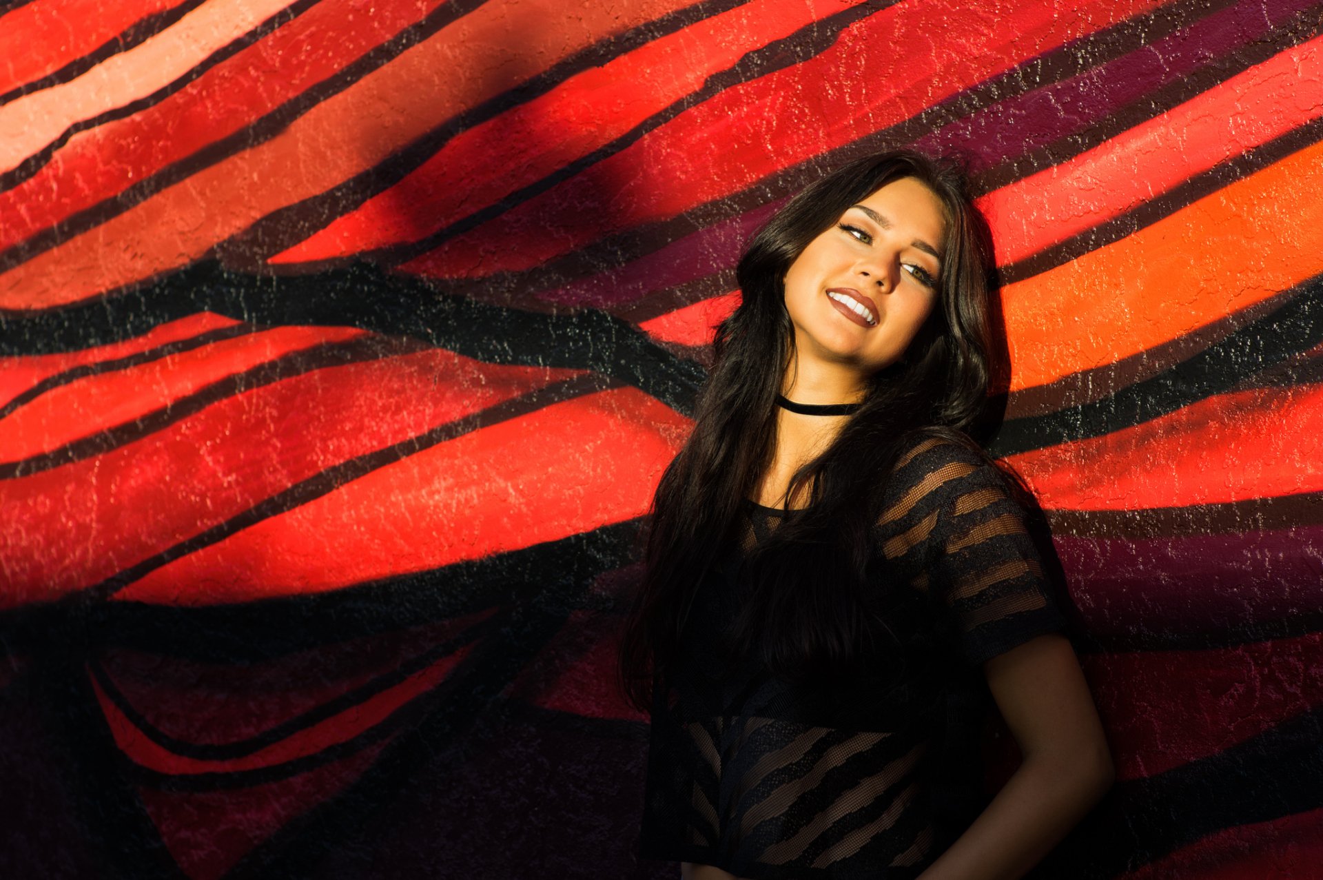 A brunette woman with green eyes and a black choker necklace poses confidently against a vibrant, red graffiti wall. This high-definition image serves as a striking desktop wallpaper background.