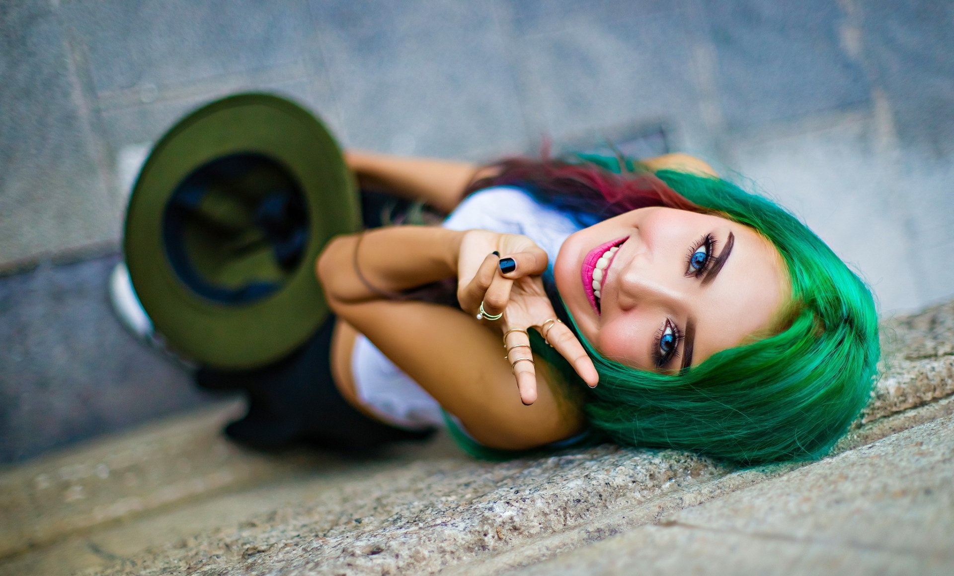 A smiling woman with vibrant green hair, bright blue eyes, and pink lipstick poses outdoors, holding a green hat, captured in HD for a desktop wallpaper background.
