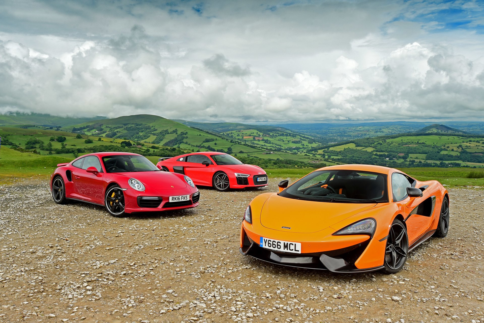 Three supercars—an orange McLaren 570S, a red Porsche 911, and an Audi R8—are parked on a gravel surface with rolling hills and cloudy skies in the background.