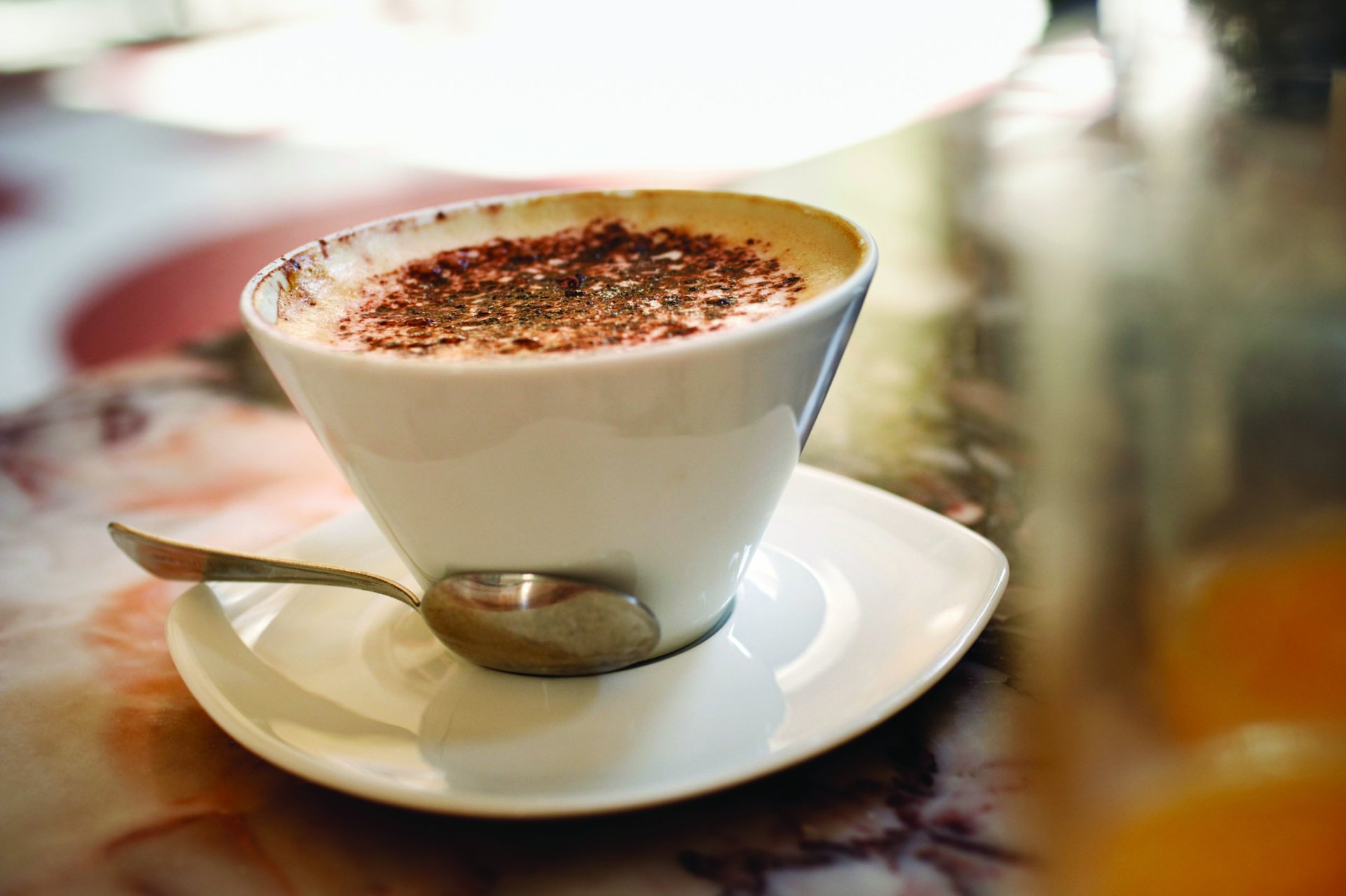 HD desktop wallpaper featuring a close-up of a white cup of coffee with sprinkled cocoa on top, placed on a saucer with a spoon, showcasing a warm food and drink moment.