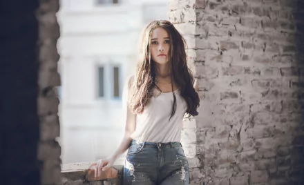 HD desktop wallpaper featuring a brunette woman model standing by a rustic brick wall, softly lit with natural light.