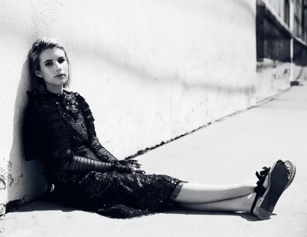 Black and white HD desktop wallpaper featuring American actress Emma Roberts seated against a wall, dressed in a dark, textured outfit.