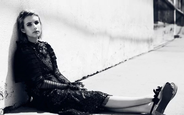 Black and white HD desktop wallpaper featuring American actress Emma Roberts seated against a wall, dressed in a dark, textured outfit.