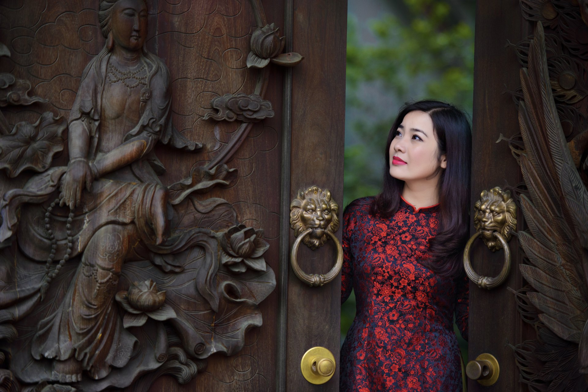 A brunette woman with brown eyes and lipstick stands elegantly at a decorative door, showcasing a beautiful dress. The image features intricate carvings, creating a stunning HD wallpaper.