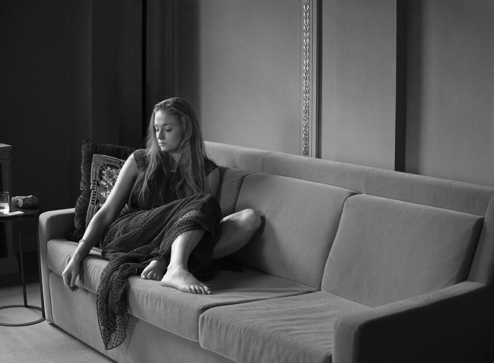 Black and white HD desktop wallpaper featuring English actress Sophie Turner seated thoughtfully on a couch in a minimalist setting.