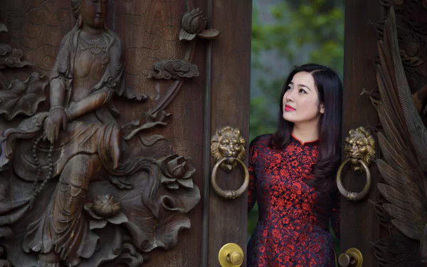 A brunette woman with brown eyes and lipstick stands elegantly at a decorative door, showcasing a beautiful dress. The image features intricate carvings, creating a stunning HD wallpaper.
