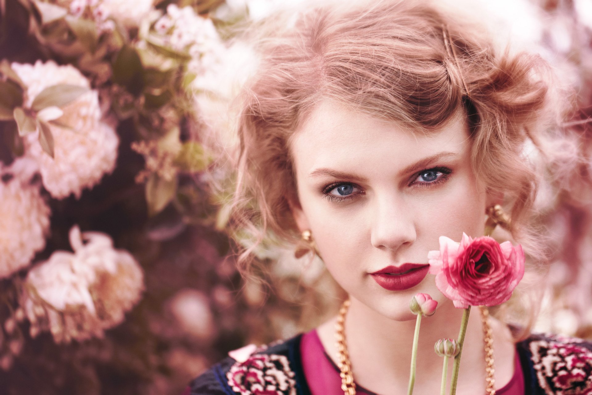 A close-up of a blonde woman with blue eyes and red lipstick, holding a flower, set against a backdrop of pink blossoms, capturing a serene and artistic vibe.