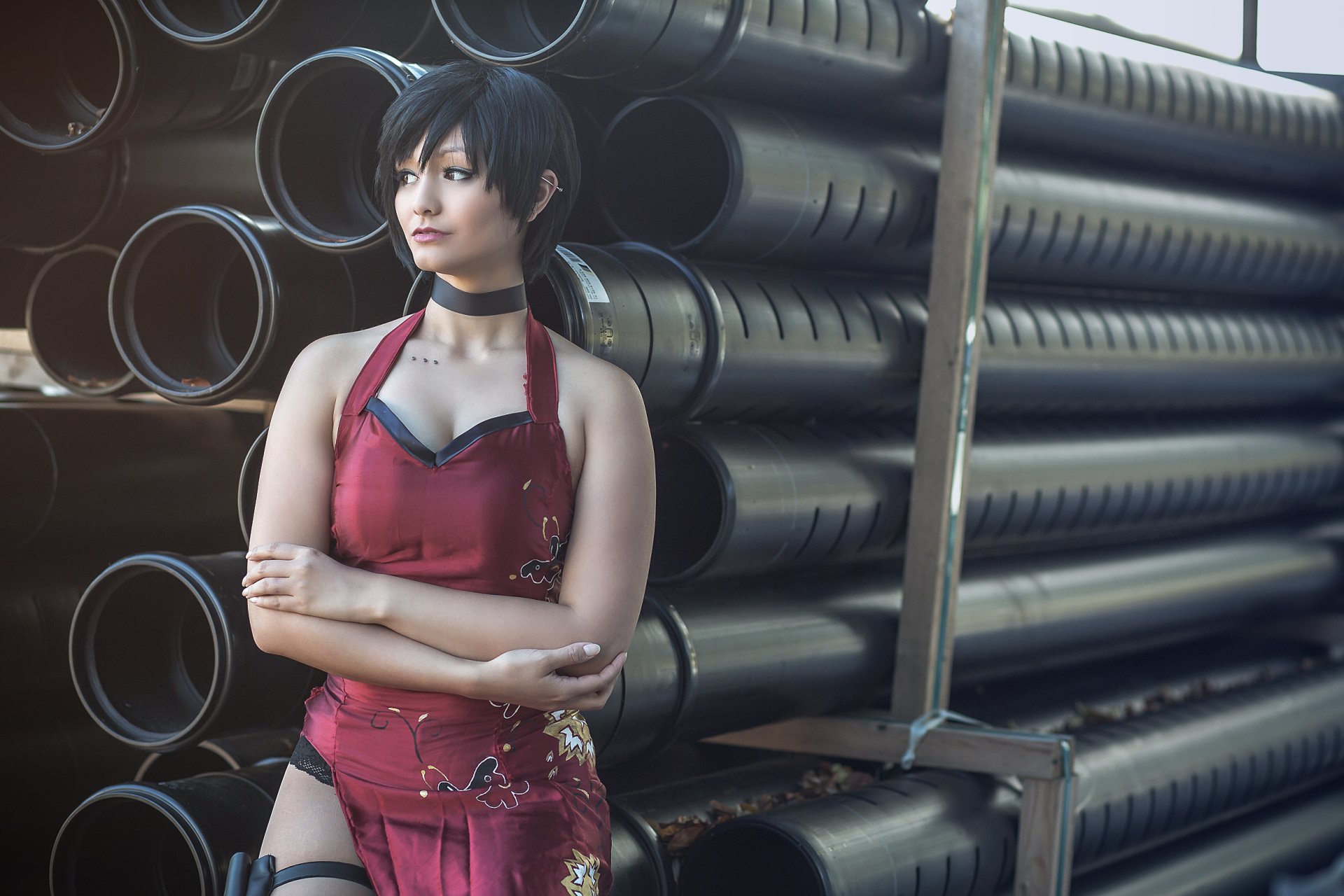 Woman with short black hair in an Ada Wong cosplay from Resident Evil stands confidently against a backdrop of industrial pipes, captured in vibrant HD detail.