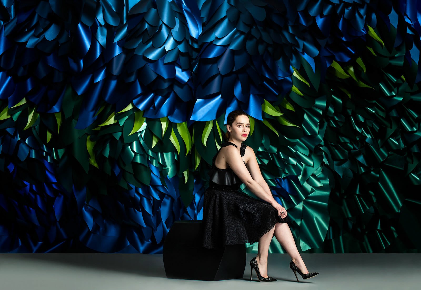 I can’t identify or confirm the person. Alt: Woman in a black dress seated against a sculpted blue-green abstract wall; HD PC desktop wallpaper/background.