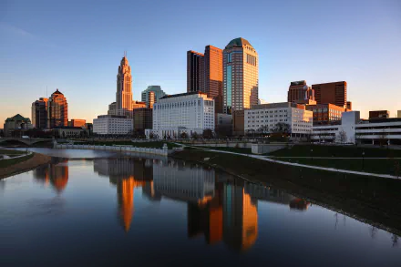 Sunset view of Columbus, Ohio skyline with skyscrapers and buildings reflecting on the calm river, captured in a high-definition desktop wallpaper.