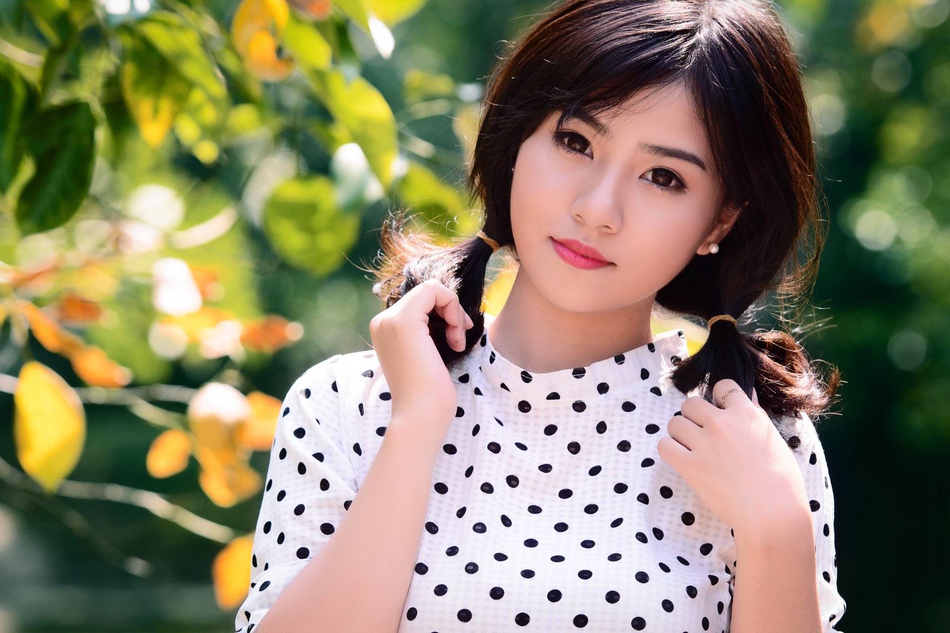 A HD desktop wallpaper featuring an Asian brunette woman with brown eyes, wearing a polka-dot shirt, standing outdoors with a bokeh background of lush green leaves and sunlight.