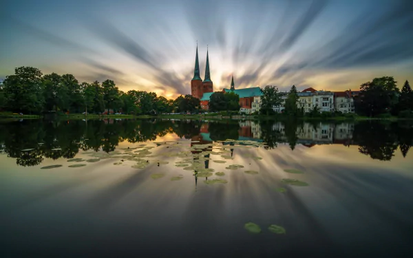 Sunset over Lübeck Cathedral: twin spires and trees mirrored in a pond under a dramatic sky in Lübeck, Germany. 2K Quad HD PC desktop wallpaper.