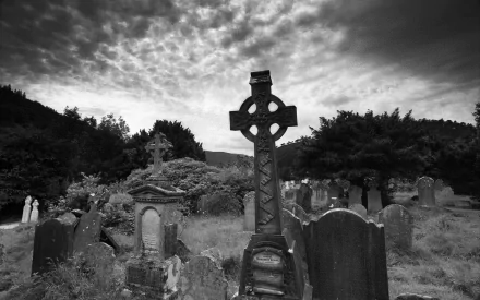 A black-and-white image of a cemetery featuring a prominent Celtic cross headstone, surrounded by weathered gravestones and a moody, cloudy sky.