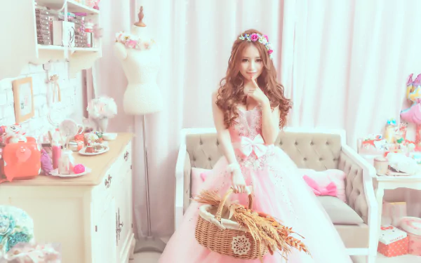 A brunette woman with brown eyes wearing a pink dress and a floral wreath stands in a pastel-colored room holding a wicker basket. The setting is elegant, making this HD image an enchanting desktop wallpaper.
