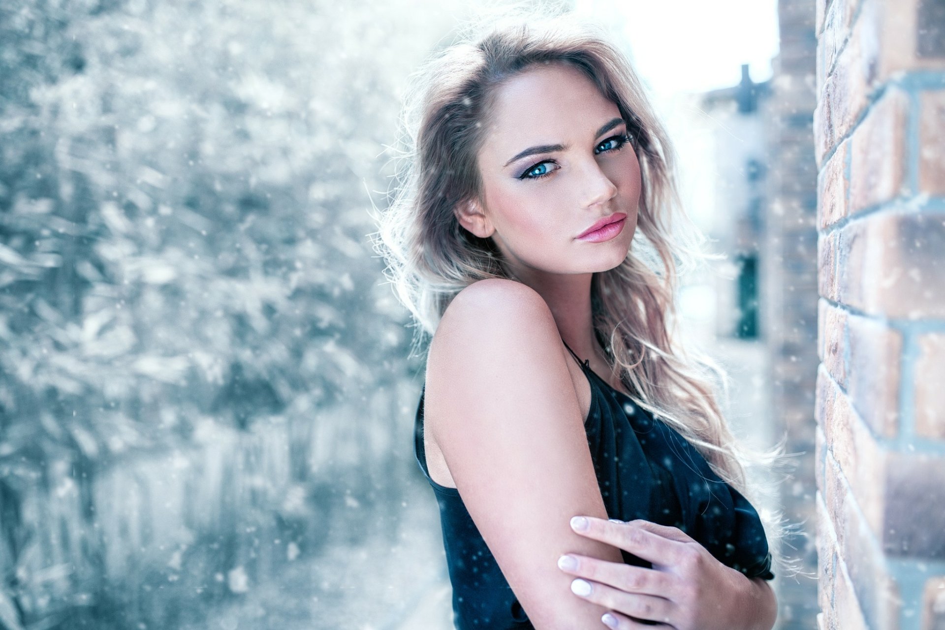 Portrait of a blonde woman with blue eyes standing against a brick wall in a snowfall. The image is a high-definition desktop wallpaper and background.