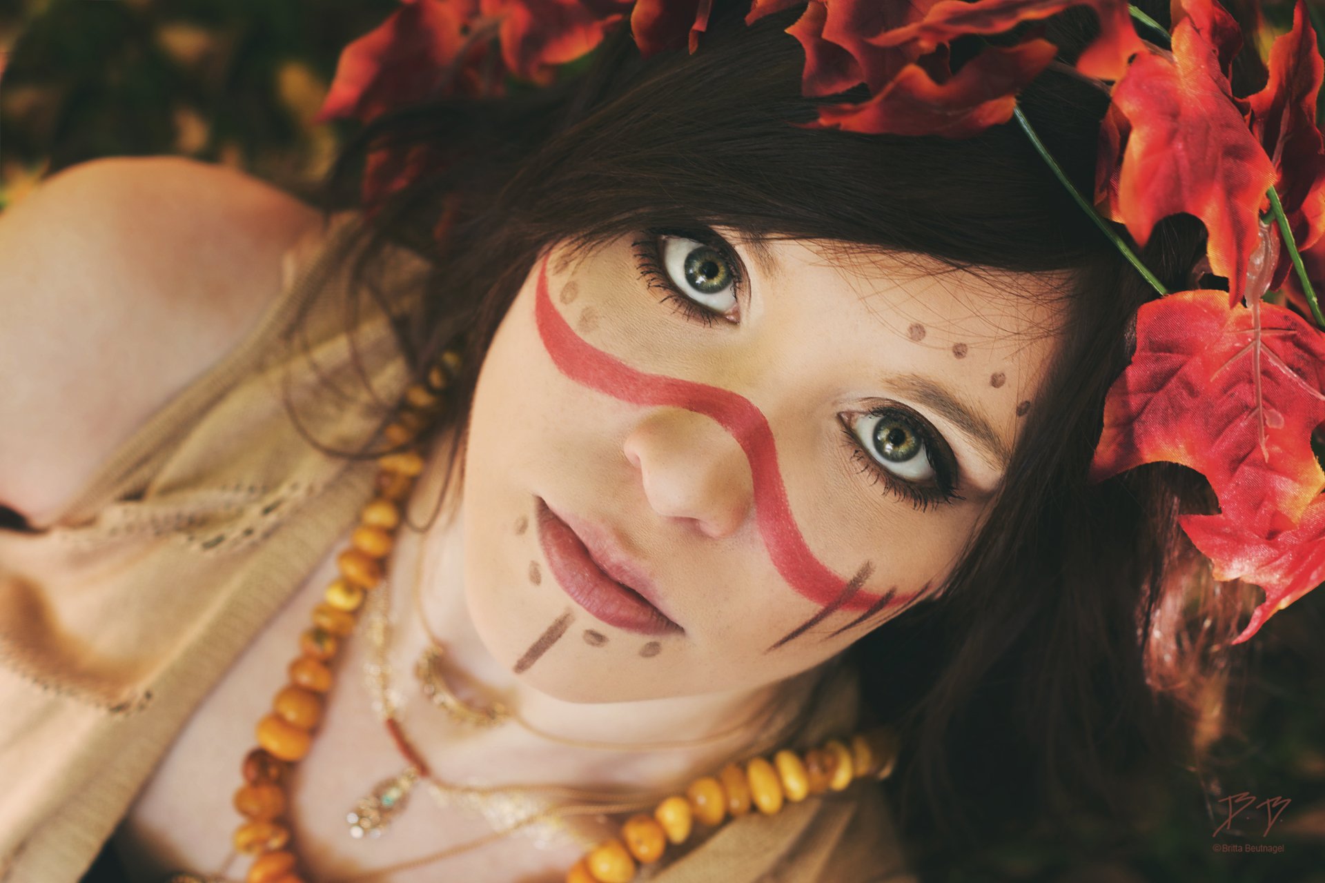 HD desktop wallpaper featuring a Native American brunette model with green eyes, intricate face makeup, and a crown of red leaves.
