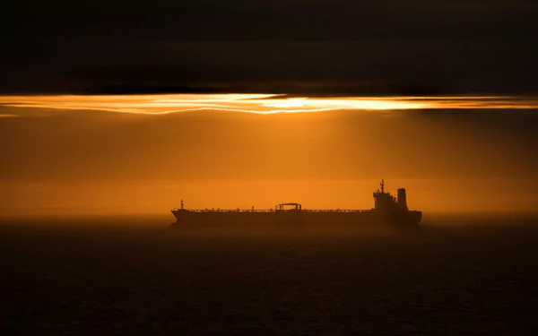 A silhouetted tanker ship sits against a vibrant sunset, casting a striking atmosphere over the sea. This HD image serves as an eye-catching desktop wallpaper and background.
