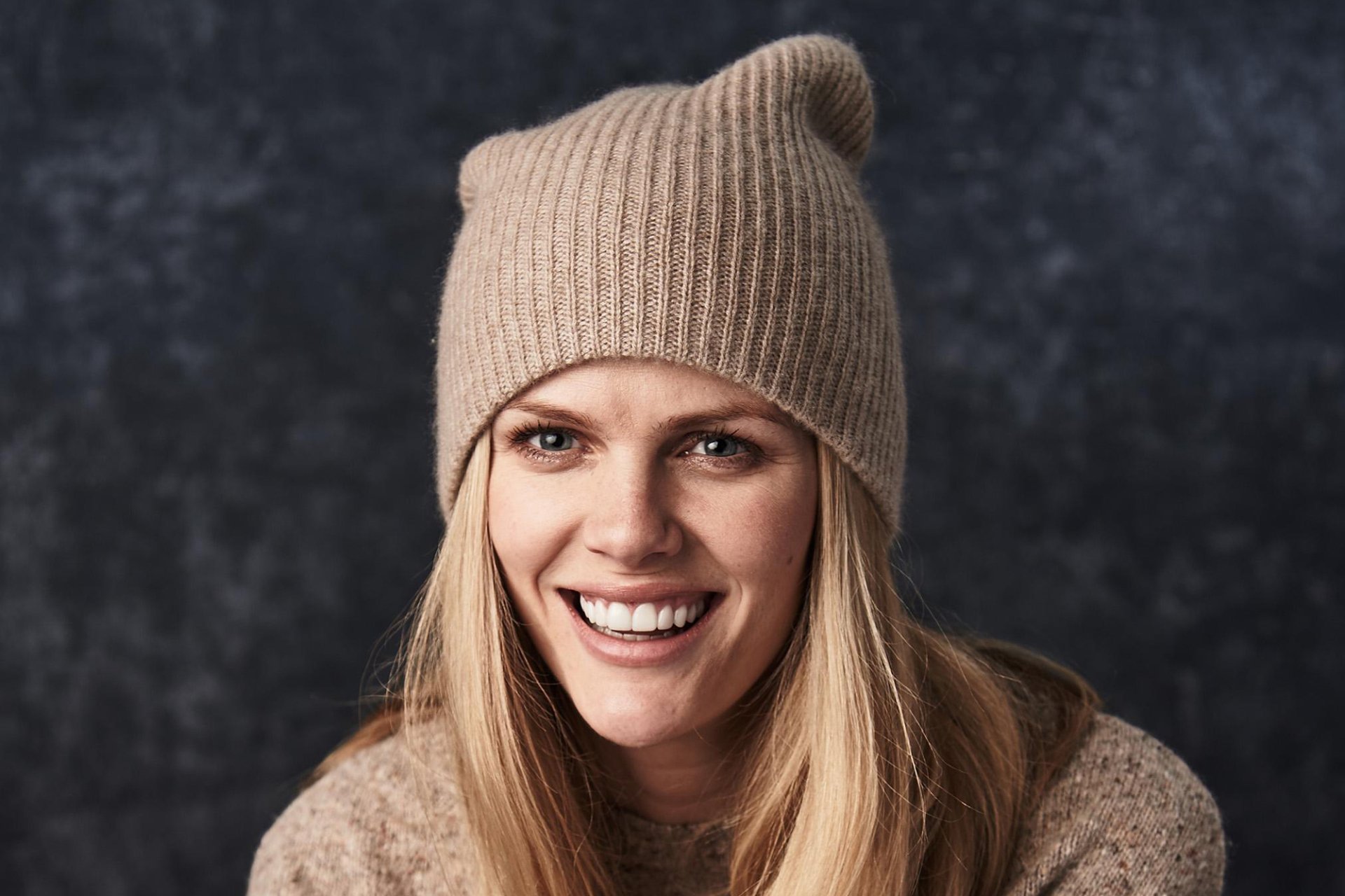 Smiling blonde American model wearing a beige knit hat and sweater, close-up portrait on a textured dark background — HD desktop wallpaper.