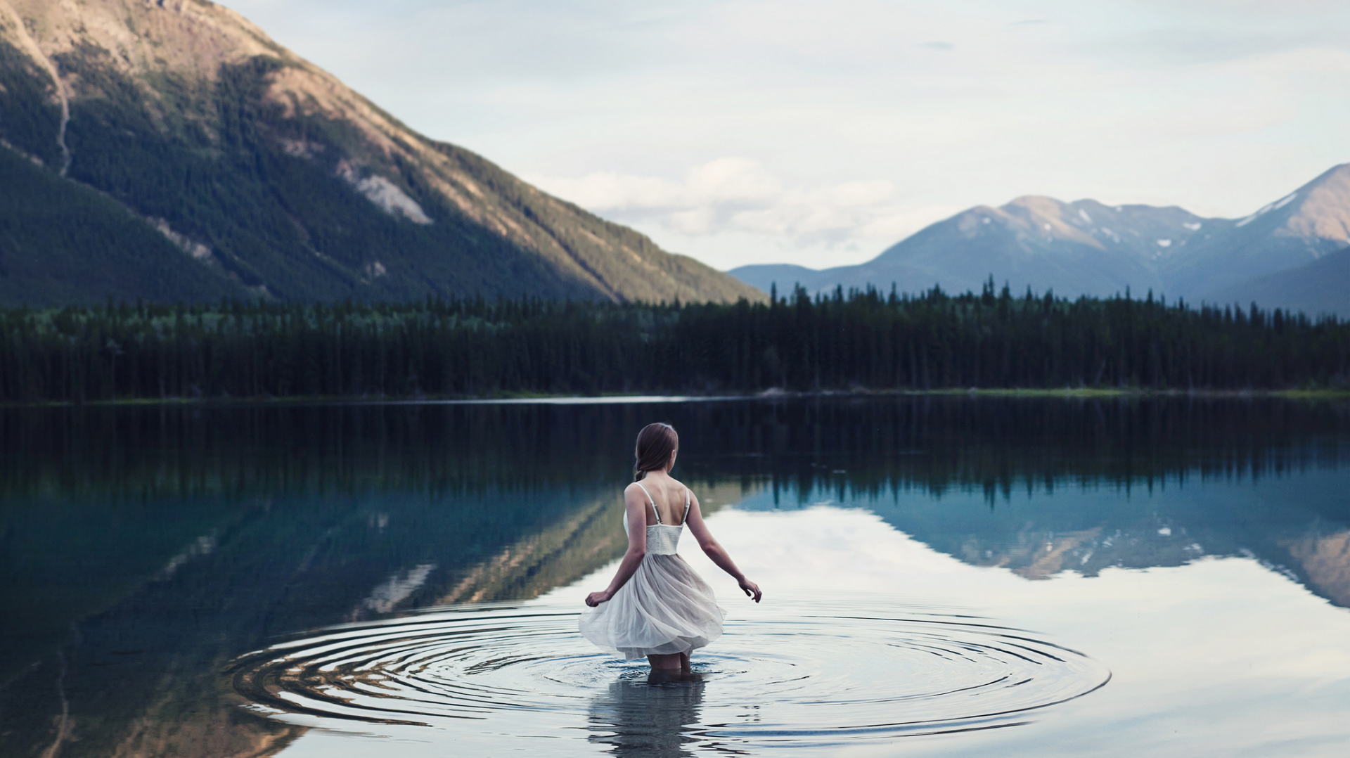HD PC desktop wallpaper: woman shown from rear wading into a calm mountain lake, white dress trailing, ripples and mirrored peaks under soft light.
