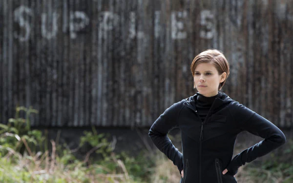 HD desktop wallpaper of a woman in a black jacket standing with hands on hips before a weathered wooden wall stenciled SUPPLIES; a still from the film Morgan (2016).