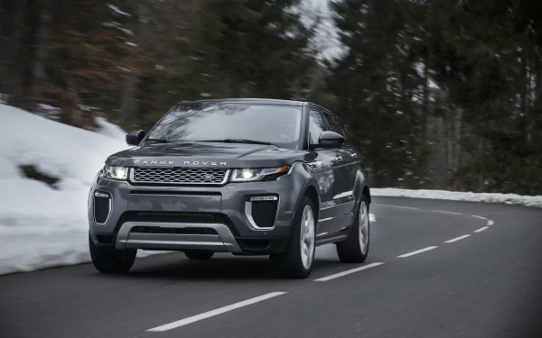 A sleek silver Land Rover Range Rover Evoque drives along a snow-lined road, showcasing its sporty design against a backdrop of lush trees. 4K Ultra HD quality enhances the visual appeal.