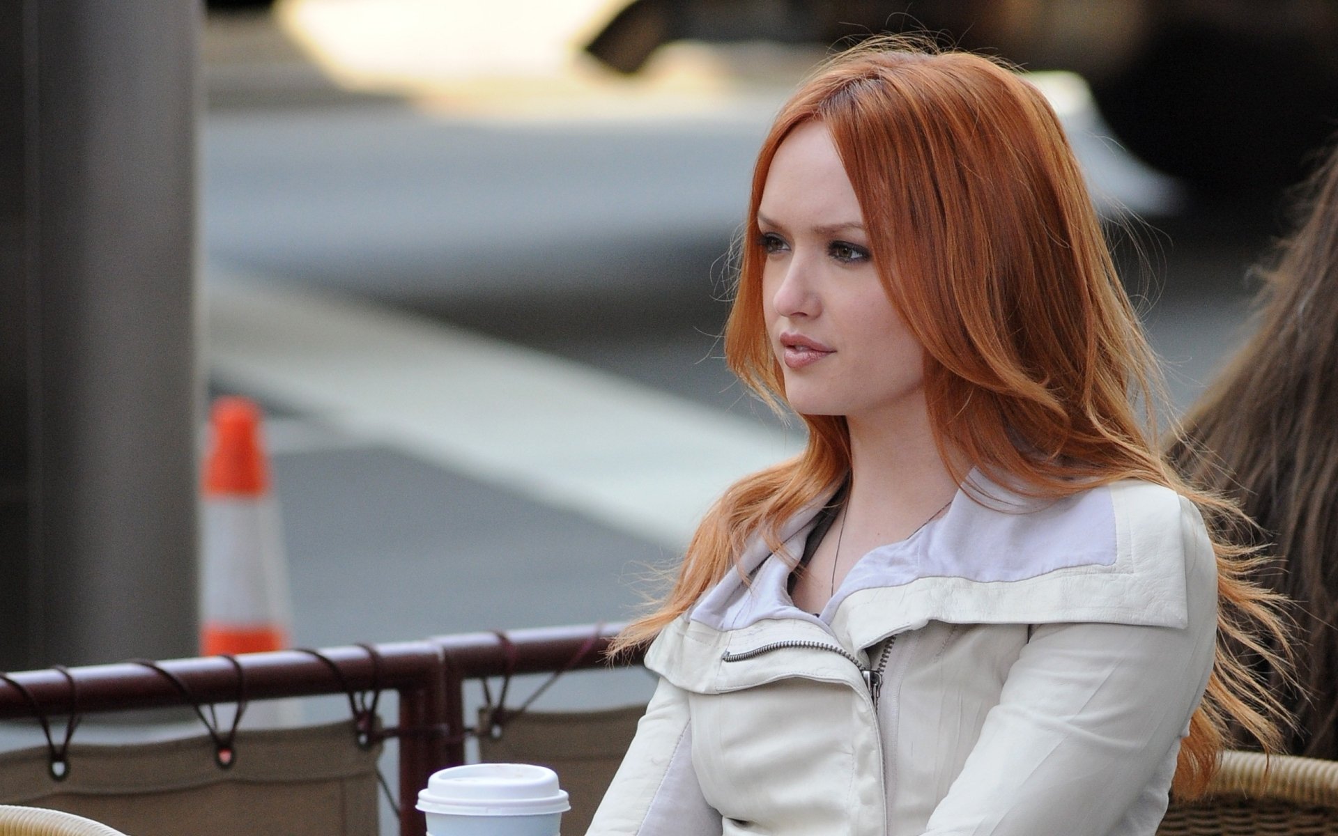 A striking woman with long red hair sits outdoors, sipping coffee, dressed in a stylish jacket. This HD image serves as a vibrant desktop wallpaper and background.