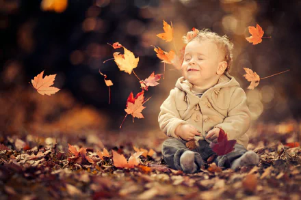 A cute little boy sits among fallen autumn leaves with bokeh background, captured in vibrant fall colors—an HD desktop wallpaper showcasing joyful childhood moments.