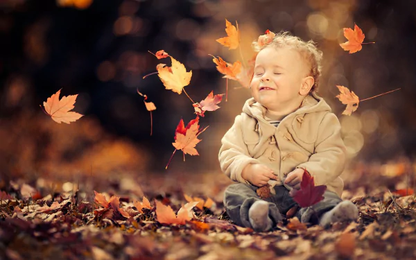 A cute little boy sits among fallen autumn leaves with bokeh background, captured in vibrant fall colors—an HD desktop wallpaper showcasing joyful childhood moments.