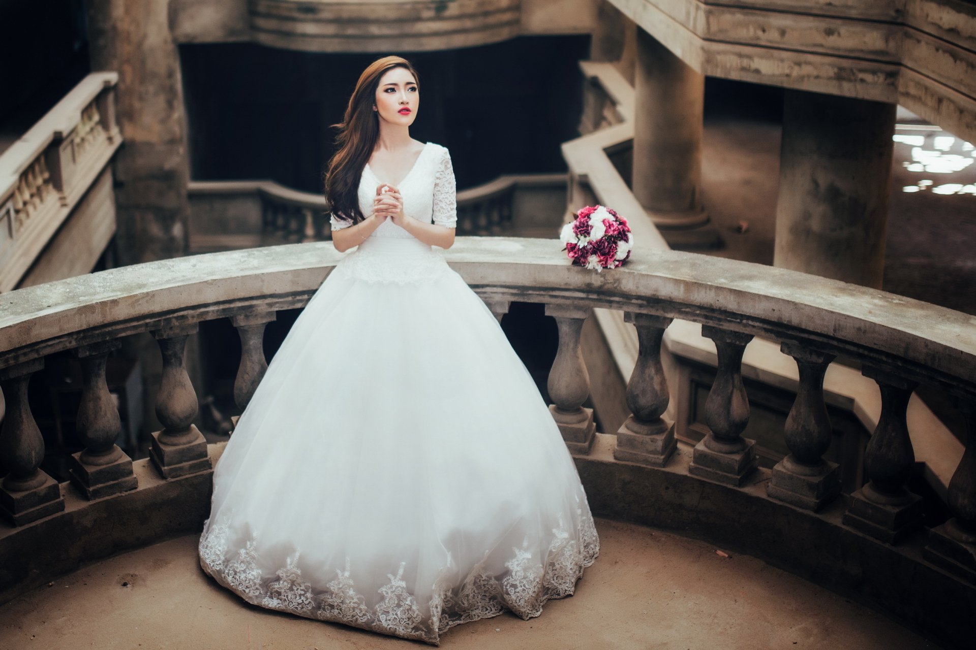 HD desktop wallpaper featuring an Asian bride with brunette hair and red lipstick, wearing a white wedding dress, standing gracefully on a stone balcony with a bouquet nearby.