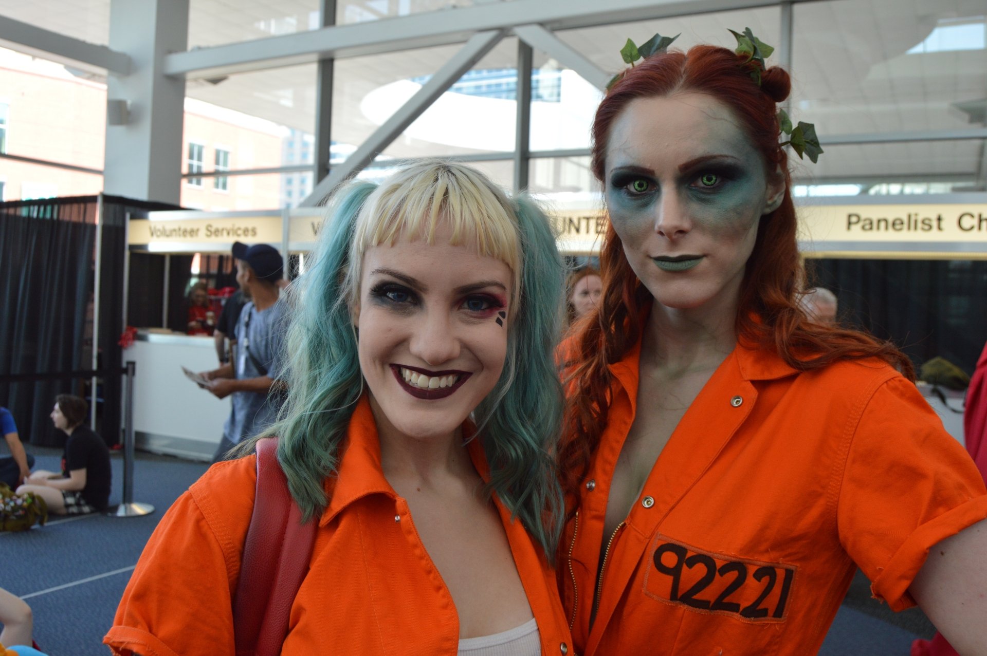 Two women cosplaying Poison Ivy and Harley Quinn in orange prison jumpsuits; Ivy with leaf crown and red hair, Harley with blue-green hair and bold makeup. 5K Ultra HD PC desktop wallpaper.