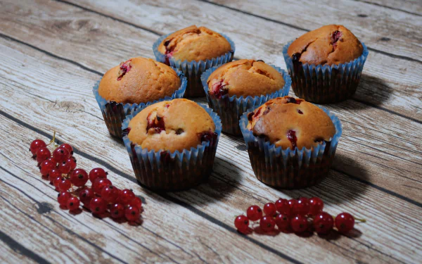Six currant muffins arranged on a wooden surface with clusters of fresh red currants, captured in a high-definition desktop wallpaper featuring dessert and pastry elements.