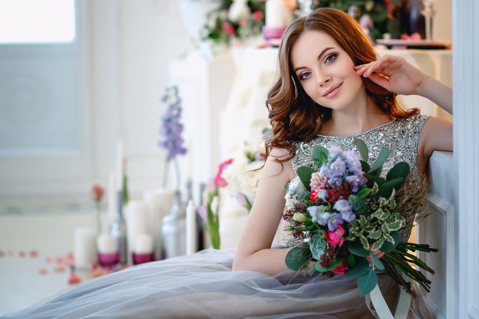 A brunette woman with blue eyes smiles while holding a bouquet. She is dressed elegantly, set against a beautifully decorated background. This HD desktop wallpaper exudes charm and grace.