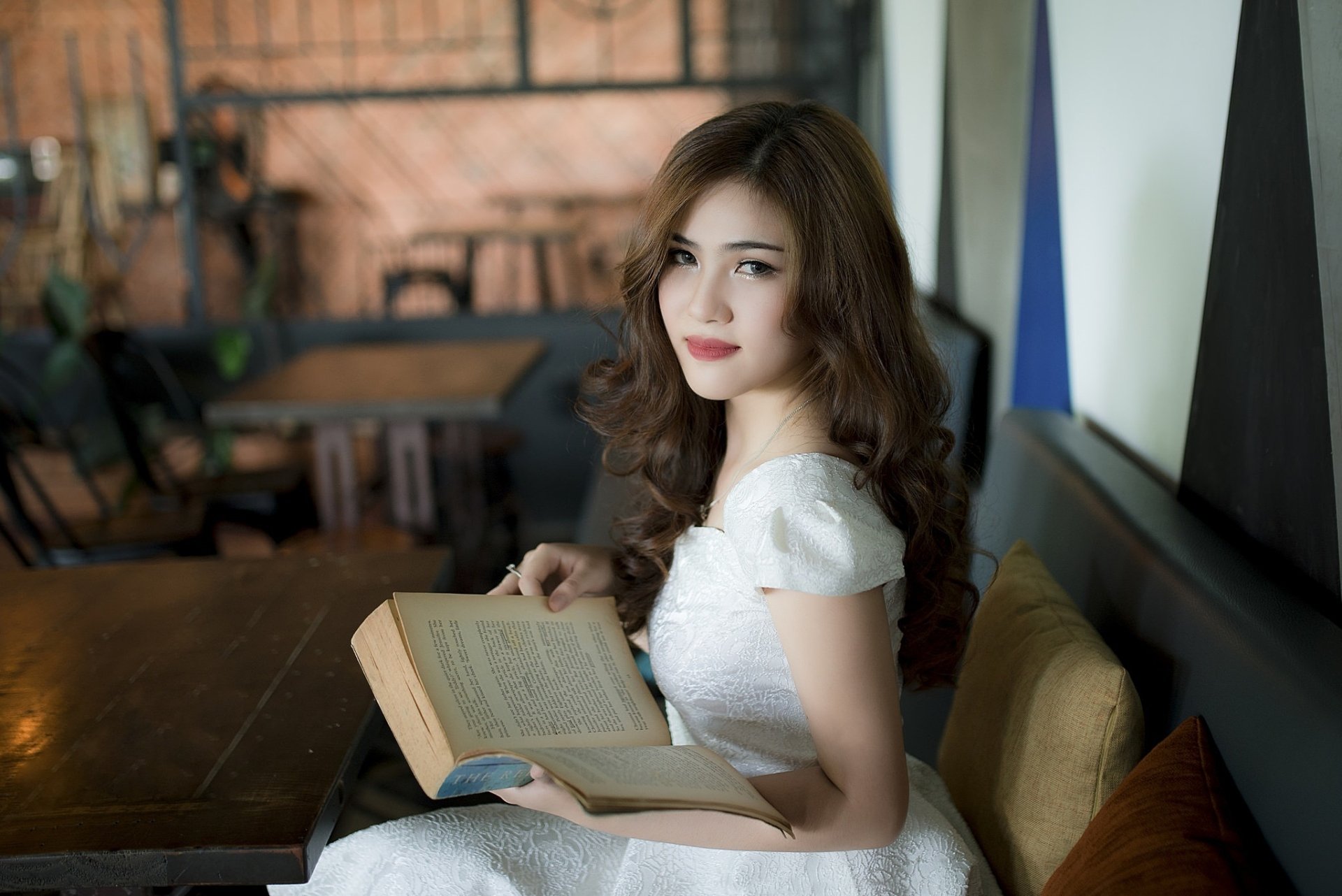 A brunette woman with brown eyes, dressed in a white dress, holding a book while seated in a cozy, softly lit room. The image is an HD desktop wallpaper and background.