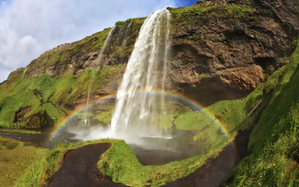 Seljalandsfoss waterfall in Iceland tumbling past mossy cliffs, a vibrant rainbow over grassy banks — 5K Ultra HD PC desktop wallpaper.