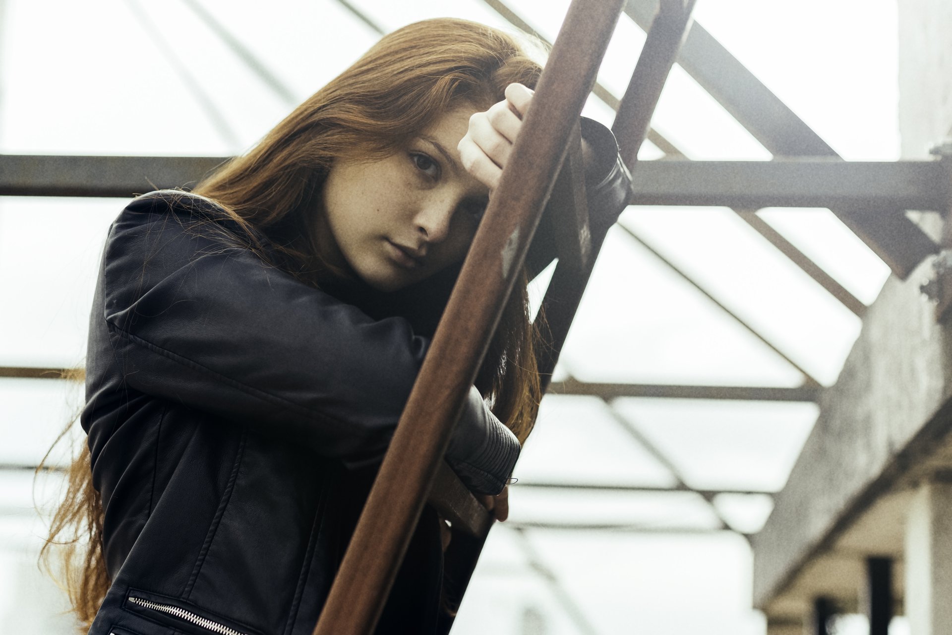 A redhead woman model in a black jacket leans on a metal railing, captured in a crisp 4K Ultra HD PC desktop wallpaper background with industrial elements.