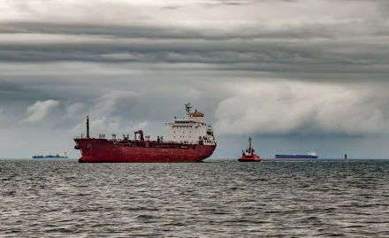 A red tanker ship navigates calm waters under a cloudy sky, accompanied by a small tugboat, creating a striking seascape for a desktop wallpaper.