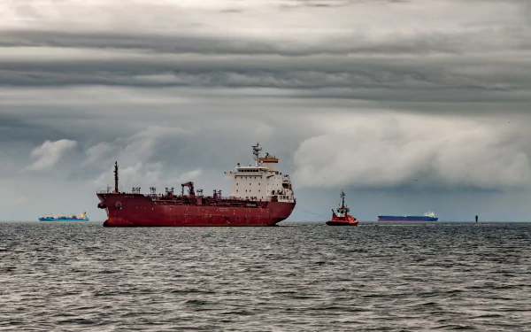 A red tanker ship navigates calm waters under a cloudy sky, accompanied by a small tugboat, creating a striking seascape for a desktop wallpaper.
