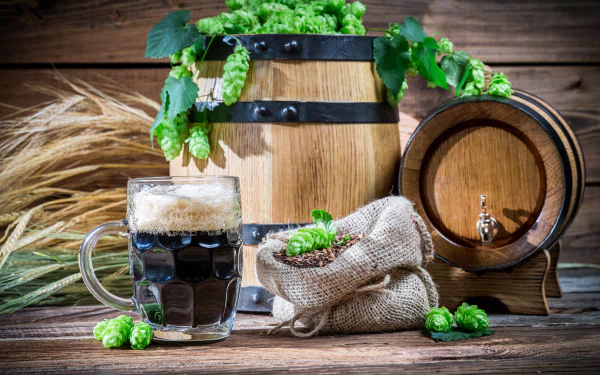 A 4K Ultra HD still life featuring a glass of dark beer, wooden barrels, fresh hops, and grains arranged on a rustic wooden surface.