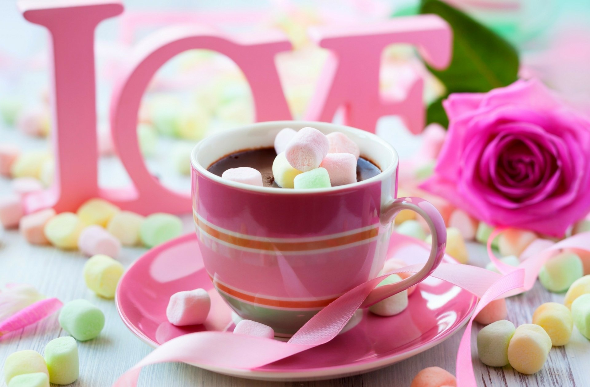 A romantic scene featuring a pink cup of chocolate adorned with marshmallows, surrounded by pastel candy and a blooming rose, set against a backdrop of the word LOVE.