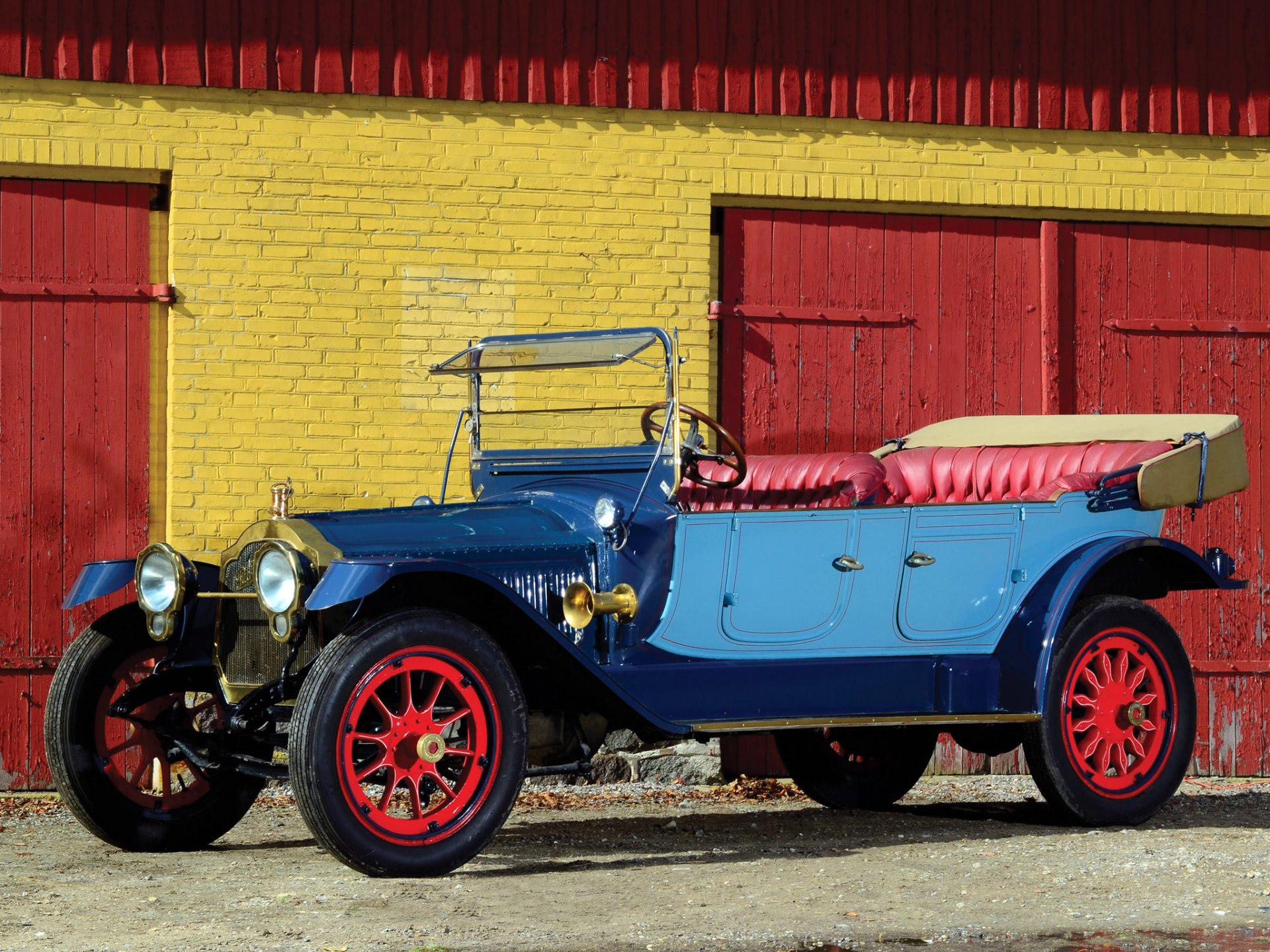 HD desktop wallpaper: blue 1914 Packard Six 7-passenger Touring vintage car with red wheels and pink interior parked against a yellow brick wall.