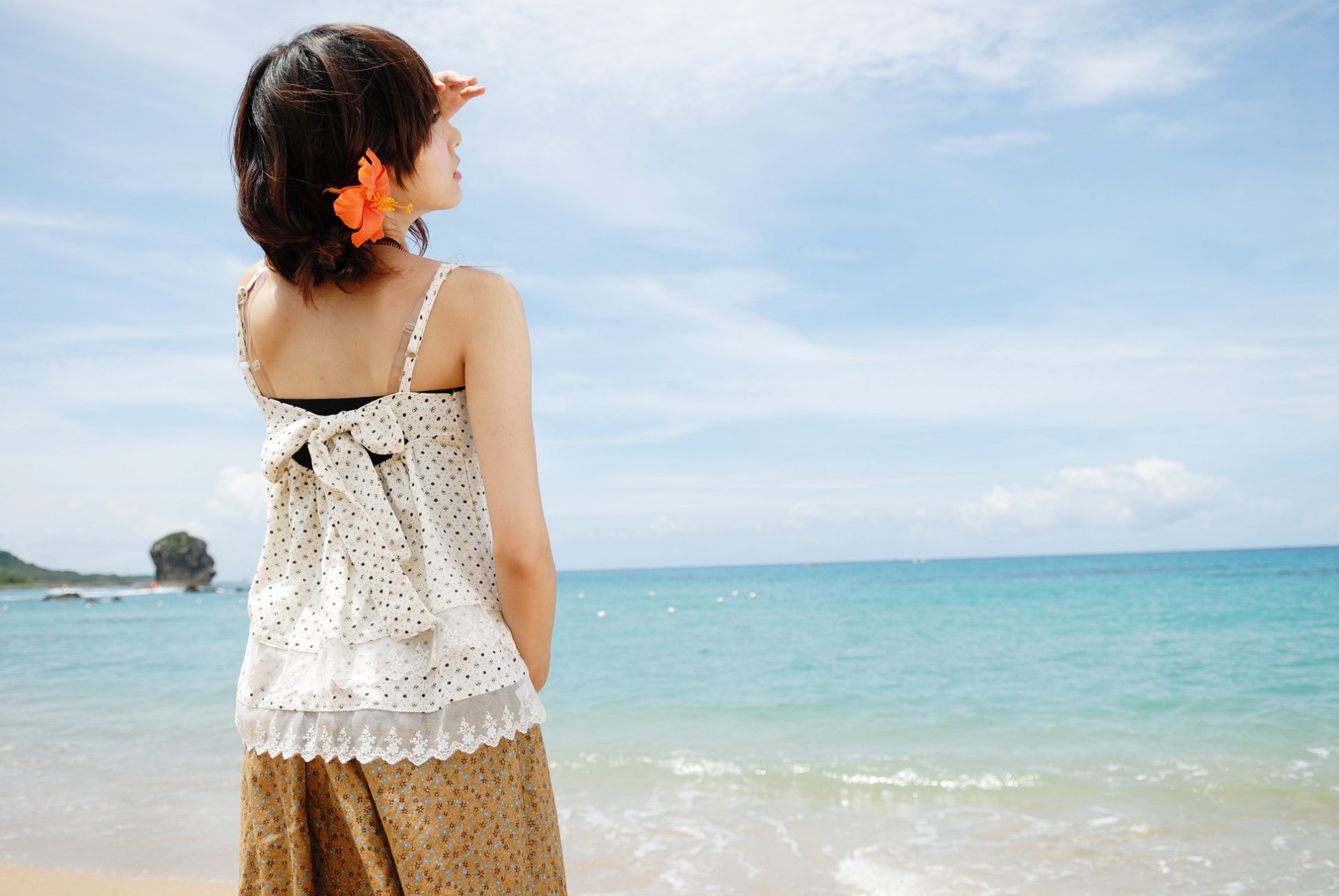 HD PC desktop wallpaper showing a woman with a flower in her hair standing on a beach, looking out at the calm blue ocean under a partly cloudy sky.