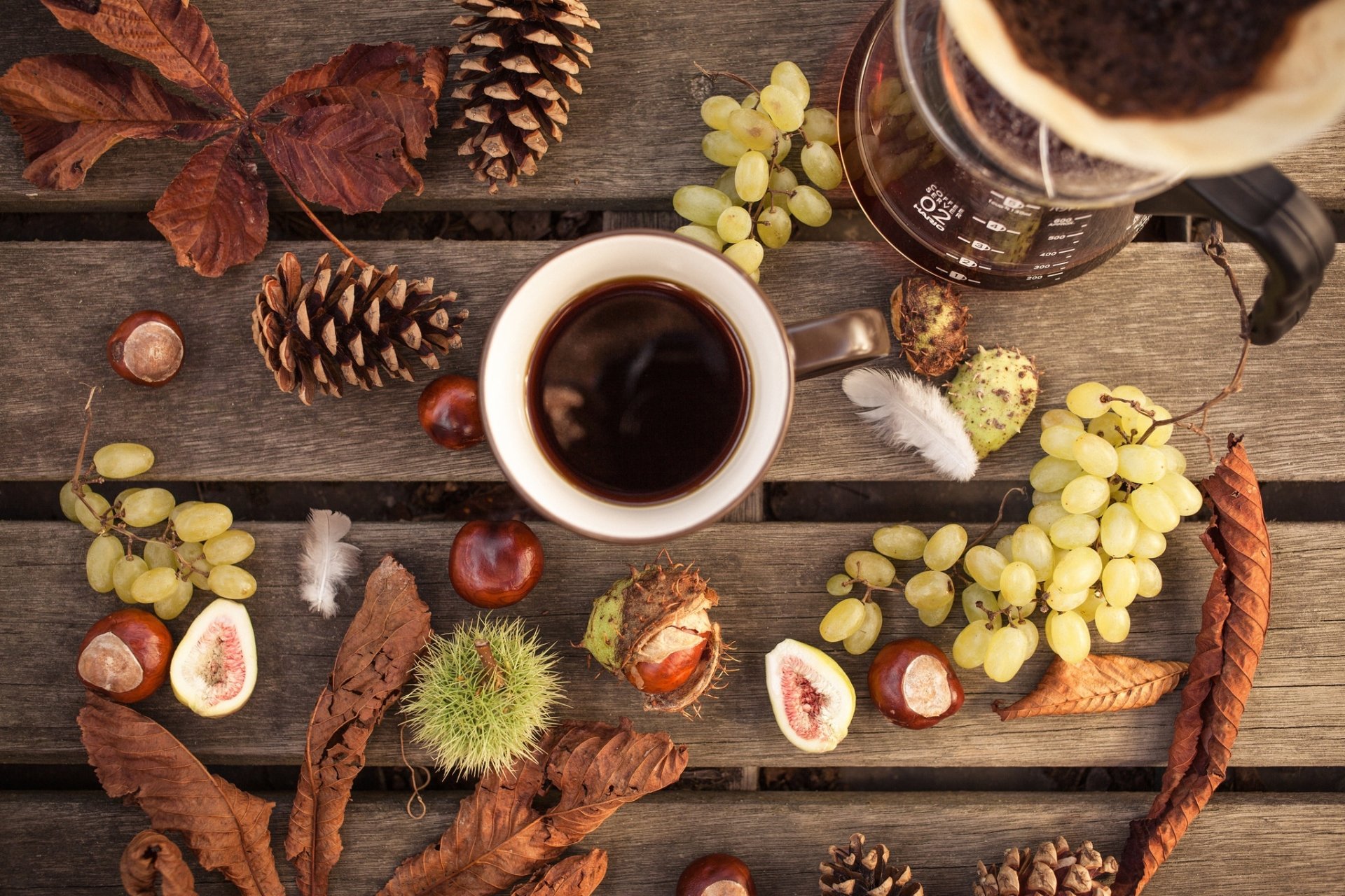 A fall-themed still life HD desktop wallpaper featuring a cup of coffee, grapes, autumn leaves, pinecones, and chestnuts arranged on a rustic wooden surface.
