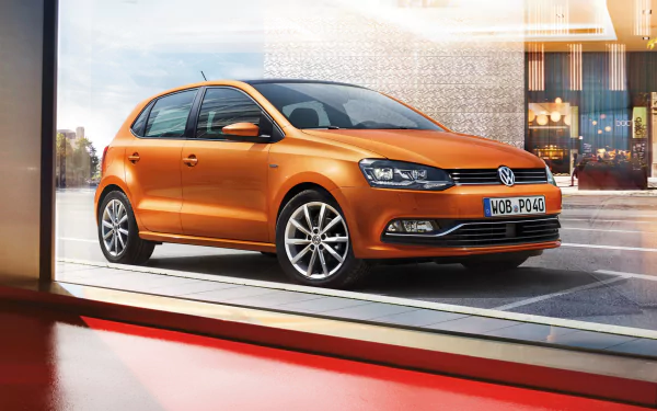 Orange Volkswagen Polo compact car in a showroom, vehicle centered against a city-lit backdrop — 4K Ultra HD PC desktop wallpaper background.