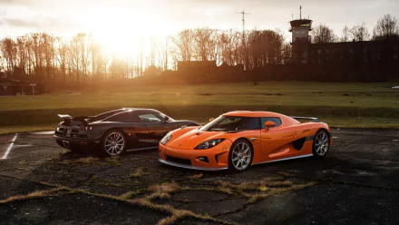 A stunning HD wallpaper featuring two Koenigsegg CCX vehicles, one black and one orange, against a scenic sunset background with a distant control tower.