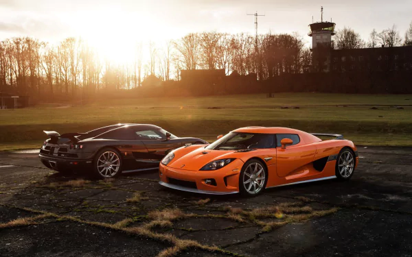 A stunning HD wallpaper featuring two Koenigsegg CCX vehicles, one black and one orange, against a scenic sunset background with a distant control tower.