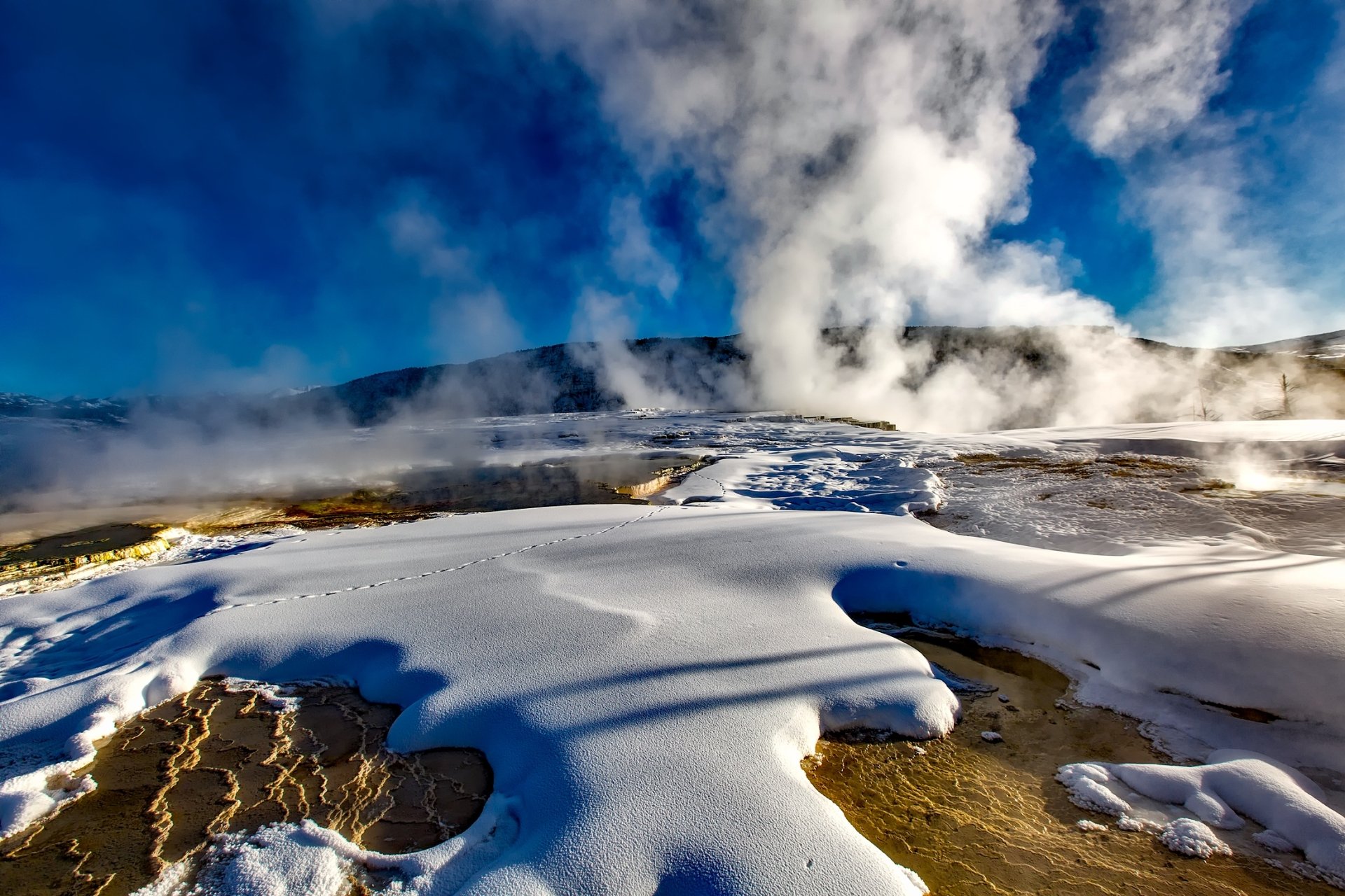 HD desktop wallpaper of a winter nature scene featuring a snowy landscape with an active geyser erupting against a vibrant blue sky.