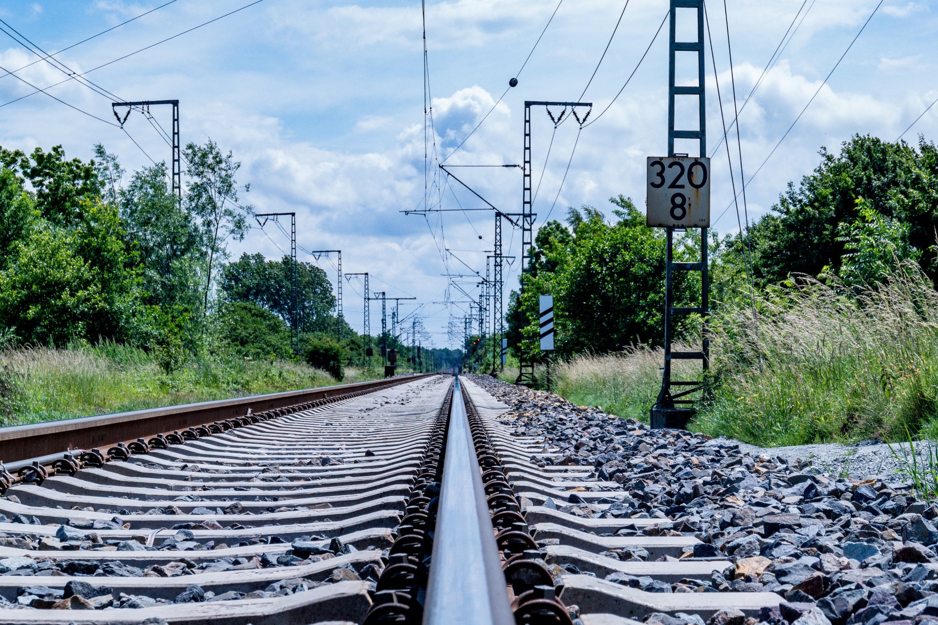 4K Ultra HD image of a man-made railroad stretching into the distance, surrounded by greenery under a partly cloudy sky, captured from ground level.